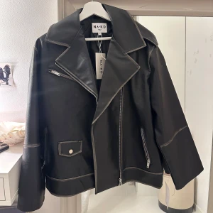 Svart skinnjacka från NA-KD - Snygg svart skinnjacka från NA-KD med dragkedja och stora slag. Jackan har långa ärmar och dekorativa sömmar som ger en cool look. Perfekt för att höja din stil till nästa nivå.