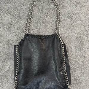 Tiamo Svart axelväska med kedjedetaljer - Snygg svart axelväska i skinn med kedjedetaljer längs kanterna. Väskan har en praktisk dragkedja på baksidan för extra förvaring. Perfekt för en stilren look.