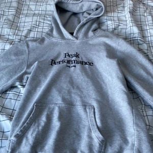 Grå hoodie från Peak Performance - Säljer en stilren grå hoodie från Peak Performance med svart logga på bröstet. Tröjan har en klassisk känguruficka och en bekväm huva. Perfekt för en avslappnad look.