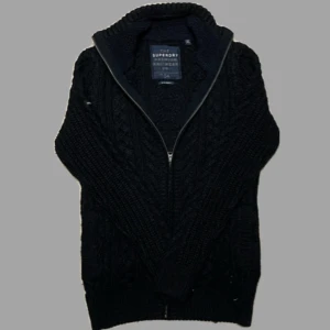 Svart stickad kofta från Superdry - Säljer en svart stickad kofta från Superdry med dragkedja framtill. Koftan har ett snyggt flätmönster och hög krage, perfekt för kyliga dagar. Den är långärmad och har ribbade muddar vid ärmslut och nederkant.