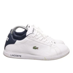 Vita sneakers från Lacoste - Snygga vita sneakers från Lacoste med mörkblå detaljer och klassisk krokodillogga på sidan. Skorna har snörning och är tillverkade i skinn, vilket ger en stilren och tidlös look. Perfekta för en avslappnad och trendig stil.