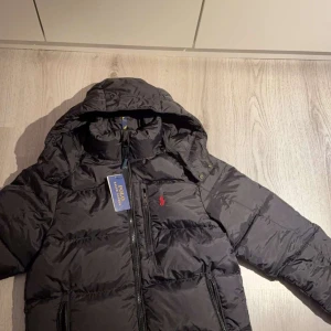 The gorham down jacket dunjacka - Polo Ralph Lauren - Helt ny svart dunjacka från Polo Ralph Lauren med huva och dragkedja. Jackan har en klassisk design med det ikoniska röda logotypbroderiet på bröstet. Perfekt för kyliga dagar. Jag fick den som födelsedagspresent men jag gillar inte den så mycket så vill hellre sälja den. Pris går att diskuteras. Dma vid minsta fundering.