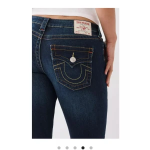 True Religion Joey Low Rise Flare Jeans - Snygga True Religion Joey jeans i mörkblå denim med låg midja och flare-stil. De har unika bakfickor med kontrastsömmar och knappar. Perfekta för en trendig look.