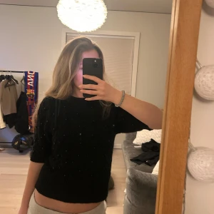 Svart tröja med pärlor från Zara - Snygg svart tröja från Zara dekorerad med små pärlor. Tröjan har en normal passform och korta ärmar, vilket gör den perfekt för både vardag och speciella tillfällen. Ett stilrent val för garderoben!