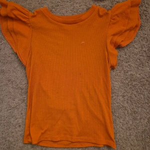 Orange ribbad topp med volangärmar - Säljer en snygg orange ribbad topp med volangdetaljer på ärmarna. Toppen har en tight passform och är perfekt för att liva upp garderoben med lite färg.