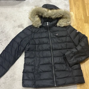 Svart dunjacka från Tommy Jeans - Snygg svart dunjacka från Tommy Jeans med en mysig fuskpäls på huvan. Jackan har en dragkedja framtill och är perfekt för kalla dagar. Den quiltade designen ger en stilren look.