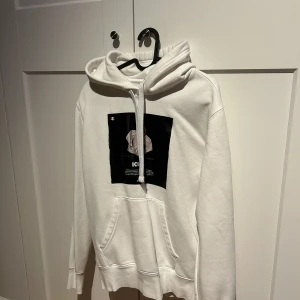 Vit hoodie från Champion - Säljer en vit hoodie från Champion med en svart tryckdetalj på framsidan. Tröjan har en klassisk känguruficka och justerbar huva med dragsko. Perfekt för en avslappnad stil.