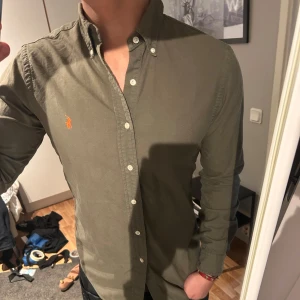 Ralph Lauren skjorta - Snygg olivgrön skjorta från Ralph Lauren med klassisk krage och knappar framtill. Skjortan har en broderad logotyp i orange på bröstet och långa ärmar. Perfekt för en stilren look.