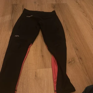 Svarta och rosa träningsbyxor från Karhu - Snygga träningsbyxor från Karhu i svart med rosa detaljer. Byxorna har dragkedjor vid bensluten och reflekterande ränder för extra synlighet. Perfekta för löpning eller gymmet.