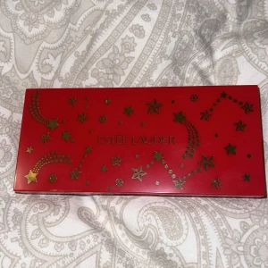 Ögonskuggspalett Celestial Glam från Estée Lauder - Snygg ögonskuggspalett från Estée Lauder i en röd förpackning med stjärndekor. Innehåller sex nyanser: Pink Flash, Aubergine Allure, Tempting Lilac, Amorous Cocoa, Nude Attitude och Smoldering Smoke. Perfekt för att skapa både vardagliga och dramatiska looks, denna ögonskuggspalett är inte använd alls.