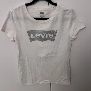 Vit t-shirt från Levi's med glittrigt tryck - Säljer en vit t-shirt från Levi's med ett glittrigt silvertryck av deras ikoniska logga på bröstet. T-shirten är kortärmad och har en klassisk rund halsringning. Perfekt för en casual look!