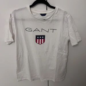 Vit t-shirt från GANT - Säljer en klassisk vit t-shirt från GANT med tryck av logotyp och ett sköldemblem i rött, vitt och blått. Perfekt för en stilren och avslappnad look. Storlek S passar M