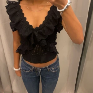 Svart volangblus - Snygg svart blus med volanger och djup v-ringning. Perfekt för en kväll ute eller en speciell tillställning. Blusen är ärmlös och har en croppad passform som ger en trendig look.