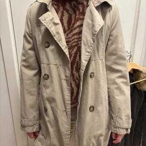 Beige trenchcoat från 365 Sunshine - Snygg beige trenchcoat med klassisk design från 365 Sun & Shine. Jackan har knappar framtill och en stilren krage. Perfekt för vår och höst med sin tidlösa look.