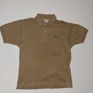 Beige pikétröja från Lacoste - Säljer en klassisk beige pikétröja från Lacoste med korta ärmar och den ikoniska krokodilloggan på bröstet.
