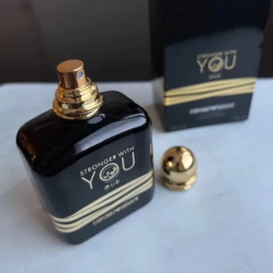 Stronger With You Oud exclusive edition - Elegant parfymflaska i mörkt glas med gyllene detaljer och en matchande gyllene kork. Doften 'Stronger With You Oud' är en lyxig och intensiv doftupplevelse. Förpackningen är stilren och svart med gyllene text och detaljer.