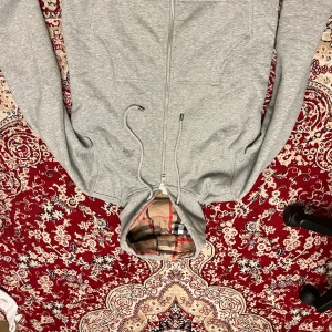 Grå hoodie med dragkedja från Burberry - Snygg grå hoodie från Burberry med klassiskt rutigt foder i huvan. Tröjan har dragkedja framtill och justerbara snören vid huvan. Perfekt för en stilren och bekväm look.