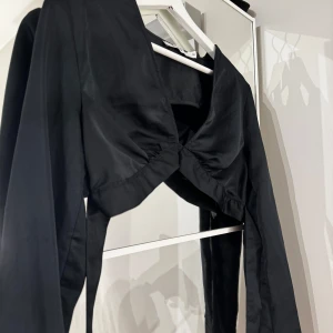 Svart långärmad topp med knyt - Snygg svart långärmad topp med knyt i ryggen. Perfekt för en stilren look med en twist. Toppen har en unik design med en öppen rygg och knytband som ger en elegant touch.