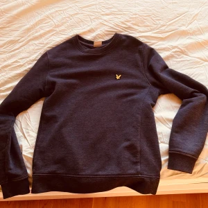 Mörkgrå tröja från Lyle & Scott - Säljer en stilren mörkgrå tröja från Lyle & Scott med deras ikoniska gula logga på bröstet. Tröjan är tillverkad i en mjuk bomulls- och polyesterblandning och har långa ärmar. Perfekt för en avslappnad look.