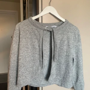 Grå stickad tröja med knytband - Mysig grå stickad tröja med knytband vid halsen. Tröjan har långa ärmar och en avslappnad passform, perfekt för kyligare dagar. Enkel och stilren design som passar till det mesta.