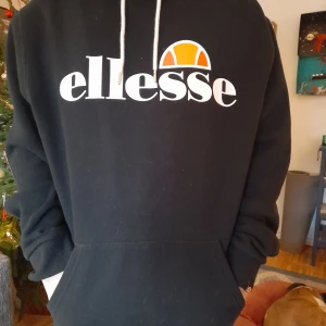 Svart hoodie från Ellesse - Säljer en svart hoodie från Ellesse med deras ikoniska logga i vitt och orange på bröstet. Hoodien har en justerbar dragsko i huvan och en stor magficka framtill. Perfekt för en avslappnad stil.