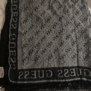 Grå och svart halsduk från Guess - Snygg och stilren halsduk från Guess i grått och svart med ett elegant mönster och logotyp längs kanten. Perfekt för att hålla dig varm och trendig under kyliga dagar. Nypris var 650kr köpt på åhles ett par år sedan.