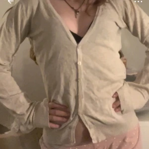 Beige kofta - Säljer en beige kofta med knappar framtill. Den har en v-ringad design och långa ärmar, perfekt för en avslappnad stil.