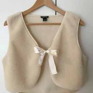 Beige fleeceväst med rosett - Superfin beige fleeceväst från Monki med en söt rosett framtill. Perfekt för lager-på-lager under hösten eller våren. Västen är v-ringad och har en mysig, mjuk känsla. Passar perfekt för en avslappnad och stilren look.