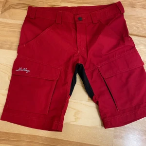 Röda shorts från Lundhags - Säljer ett par röda Lundhags shorts i storlek 36. De är perfekta för utomhusaktiviteter med sin robusta design och praktiska fickor. Shortsen har en snygg kontrast med svarta detaljer baktill. Perfekta för sommarens äventyr!