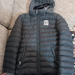 Svart dunjacka från Vinson Polo Club - Säljer en svart dunjacka från Vinson Polo Club i storlek 16. Jackan har en dragkedja framtill och en mysig huva. Perfekt för kyliga dagar med sin quiltade design. Den har också två praktiska fickor med dragkedja. Passar både till vardags och lite finare tillfällen.