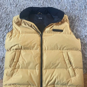 Gul dunväst från Boss - Säljer en snygg gul dunväst från Boss i storlek L. Västen är gjord av polyester och har en dragkedja framtill. Perfekt för höst och vår, med en stilren design och bekväm passform. Den har praktiska fickor och en kontrasterande svart insida. Passar både till vardags och lite finare tillfällen.