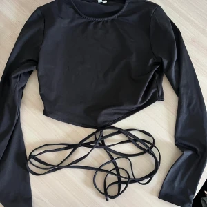 Snygg festtop med knytdetaljer  - Snygg svart topp med knytdetaljer som går 2 varv runt midjan ungefär! Använd en gång. Fint material, nästan glansigt. 