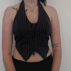 Svart randig topp - Snygg svart topp med vita ränder och knytning i nacken. Västen har knappar framtill och en stilren design som passar perfekt för festliga tillfällen. Den är croppad och ger en modern look.