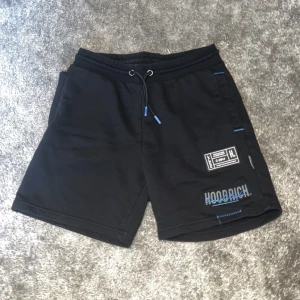 Svarta shorts från Hoodrich - Säljer ett par svarta shorts från Hoodrich i nyskick. De har en loose passform och är perfekta för sommarens alla äventyr. Shortsen har en elastisk midja med dragsko och coola blå detaljer. Perfekta för både träning och chill!
