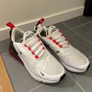 Nike Air Max sneakers - Säljer ett par Nike 270  i vitt med svarta och röda detaljer. Skorna har en stor, synlig luftkudde i hälen för extra komfort och stil. De har snörning och en modern design som passar perfekt för både träning och vardagsbruk. Använt skick