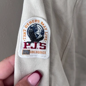 Beige hoodie från Parajumpers - Säljer en snygg beige hoodie från Parajumpers. Den har en stor logga på bröstet och en mindre patch på ärmen. Hoodien är långärmad med en justerbar huva och ribbade muddar. Perfekt för en avslappnad stil under höst och vår. Den har smink fläckar som jag inte orkat försöka ta bort där av billigt pris!