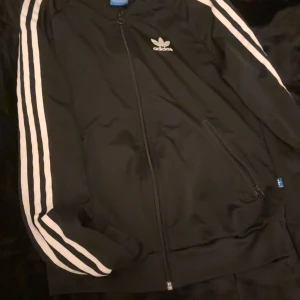 Svart träningsjacka från Adidas - Säljer en svart Adidas träningsjacka i storlek XS. Den har de klassiska vita ränderna längs ärmarna och en dragkedja framtill. Perfekt för träning eller som en snygg vardagsjacka. Adidas-loggan syns både fram och bak. Skön och stilren design!