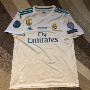 Vit Real Madrid fotbollströja och shorts - Säljer en vit Real Madrid fotbollströja med korta ärmar och matchande shorts. Tröjan har 'Fly Emirates' tryckt på framsidan och 'Ronaldo 7' på baksidan. Den är från Final Kyiv 2018 och har flera märken och logotyper. Perfekt för fans! 🏆