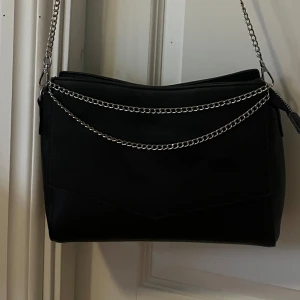 Svart axelväska från Banned Alternative - Snygg svart axelväska i syntetmaterial från Banned Alternative. Väskan har en stilren design med en kedja som axelrem och dragkedja upptill. Perfekt för både vardag och fest!