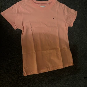 Ljusrosa t-shirt från Tommy Jeans - Säljer en snygg persikofärgad t-shirt från Tommy Jeans i storlek XS. Den är i bomull och har en liten logga på bröstet. Perfekt för sommardagar eller som ett basplagg i garderoben. Kortärmad och i nyskick!
