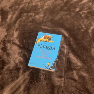 Narnia: Häxan och lejonet - Säljer boken 'Narnia: Häxan och lejonet' av C.S. Lewis. Den har ett blått omslag med en illustration av ett lejon och en lyktstolpe. Perfekt för fantasyälskare som vill dyka in i en magisk värld!