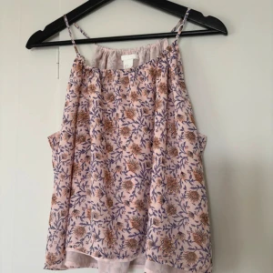 Blommig ärmlös topp - Säljer en söt blommig ärmlös topp i ljusrosa med lila och orange blommönster. Perfekt för sommardagar eller en avslappnad vårlook. Toppen har tunna axelband och en luftig passform. Passar bra till jeans eller kjol för en fräsch stil! 🌸
