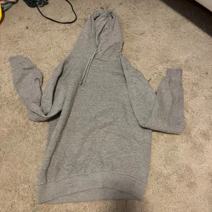 Grå hoodie från Smog - Säljer en grå hoodie från Smog i storlek M. Den är supermjuk och perfekt för kyliga dagar. Hoodien har en klassisk design med dragsko i huvan och ribbade muddar vid ärmslut och nederkant. Passformen är regular, vilket gör den bekväm och lätt att bära till vardags.