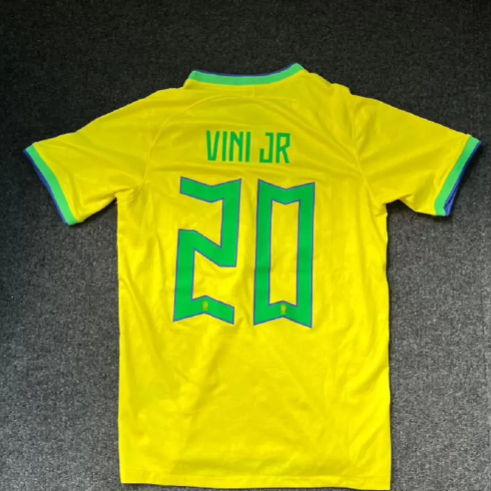 Säljer en gul Brasilien fotbollströja från Nike med nummer 20 och namnet 'VINI JR' på ryggen. Tröjan har korta ärmar och en snygg krage i grönt och blått. Perfekt för fotbollsfans som vill visa sitt stöd för Brasilien!. T-shirts.