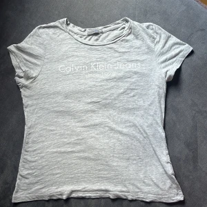 Grå t-shirt från Calvin Klein Jeans - Säljer en snygg grå t-shirt från Calvin Klein Jeans. Den är i storlek M och har en klassisk passform med korta ärmar. T-shirten är gjord av mjuk bomull och har en stilren logga på framsidan. Perfekt för en avslappnad vardagslook!