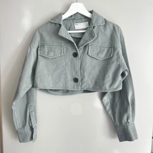 Grå croppad jacka från ASOS DESIGN - Säljer en snygg khaki croppad jacka från ASOS DESIGN. Jackan har en cool, avslappnad stil med två stora knappar framtill och två bröstfickor. oanvänd 