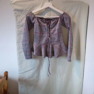 Rutig offshoulder topp med snörning - Säljer en superfin rutig offshoulder topp med snörning framtill. Den har långa ärmar och en volangdetalj vid midjan som ger en söt touch. Perfekt för höst och vår, och kan stylas både upp och ner beroende på tillfälle.