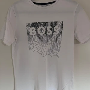 Vit t-shirt från BOSS med grafiskt tryck - Säljer en snygg vit t-shirt från BOSS i slim fit. Den har ett coolt grafiskt tryck på framsidan med texten 'BOSS' i stora bokstäver. Perfekt för en stilren och modern look! Djur- och rökfritt hem