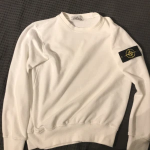 Vit tröja från Stone Island - Säljer en stilren vit tröja från Stone Island. Den har en klassisk design med långärmade ärmar och en rund halsringning. Tröjan är i bomull och har en liten fläck på baksidan men den är väldigt liten och borde gå bort i tvätten . Perfekt för både vardag och fest!