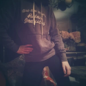 Vintage brun hoodie med tryck - Säljer en brun hoodie med texten 'Dist-1014 Mfg Co. Retail Industry' tryckt på framsidan. Den har en klassisk känguruficka och vita dragsnören vid huvan. Perfekt för en avslappnad stil!